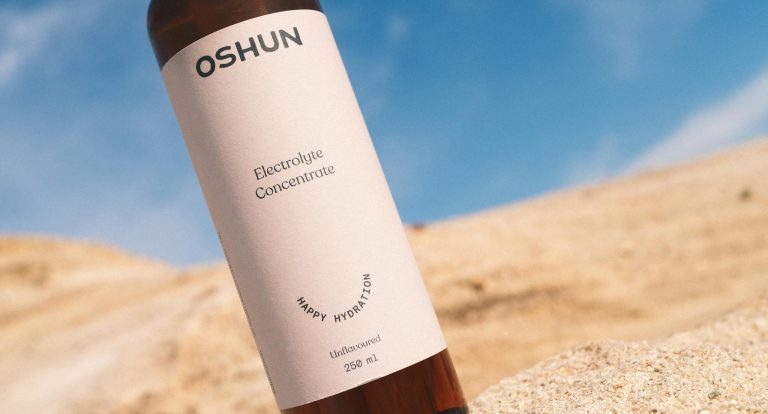 Close-up of Oshun electrolyte concentrate bottle on sand against a clear sky.